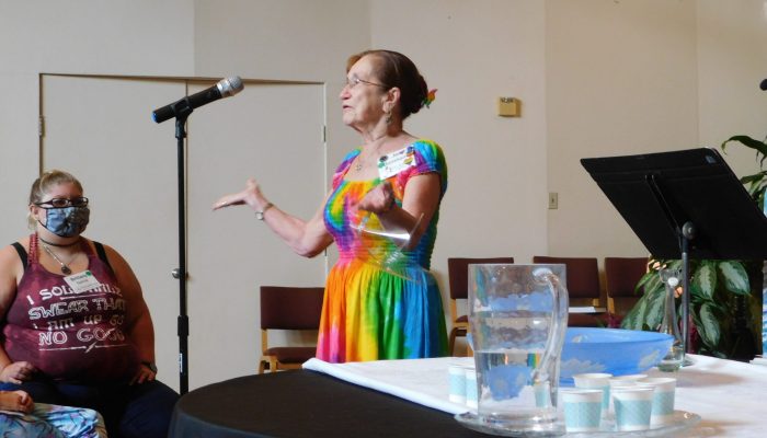 Member in rainbow dress at microphone next to a pitcher of water.